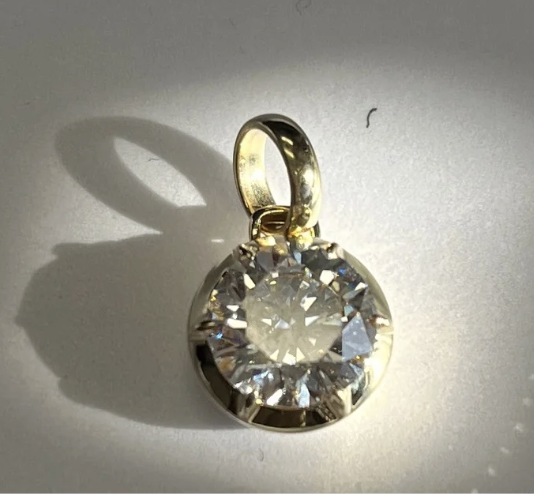The Round Lab Diamond Sola Charm features a round, clear lab-grown diamond set in a 14KT gold frame, casting a delicate shadow on a white surface under bright light.