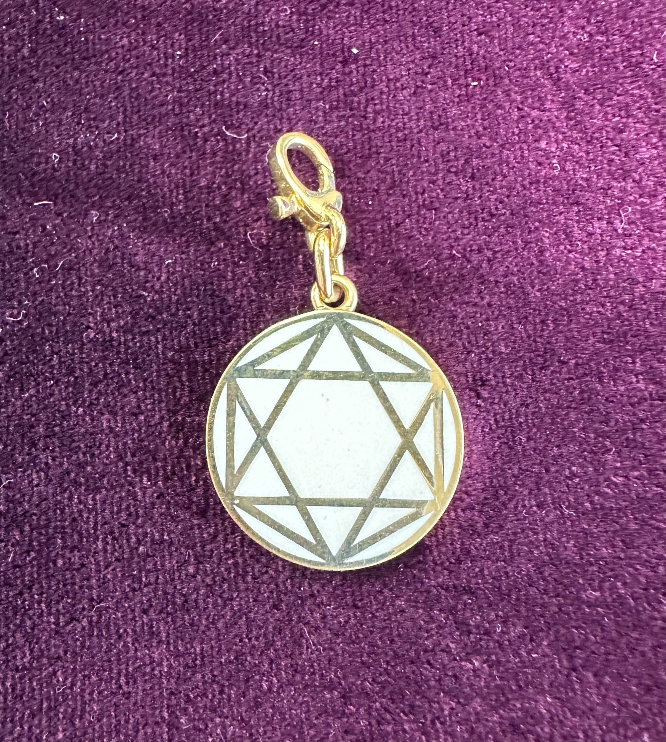 A round gold pendant featuring a geometric star pattern in white enamel, attached to a small clasp and displayed on textured purple fabric—a refined example of the White Enamel Star Charm from Janet Heller Fine Jewelry’s collection.