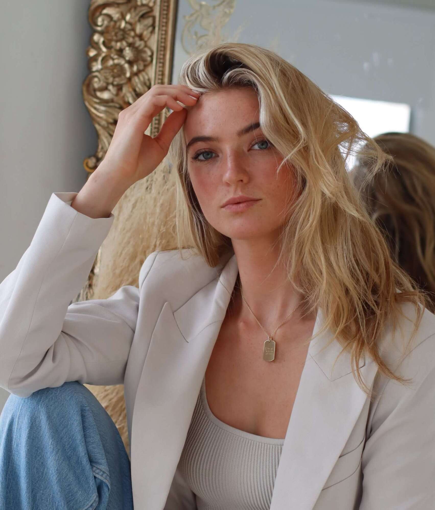 A woman with long blonde hair, wearing a white blazer and ribbed tank top, sits in front of a large ornate mirror, looking at the camera with a relaxed expression. Around her neck, she wears the Large Dog Tag Engravable Pendant Necklace—a personalized accessory that adds a meaningful touch to her elegant style.