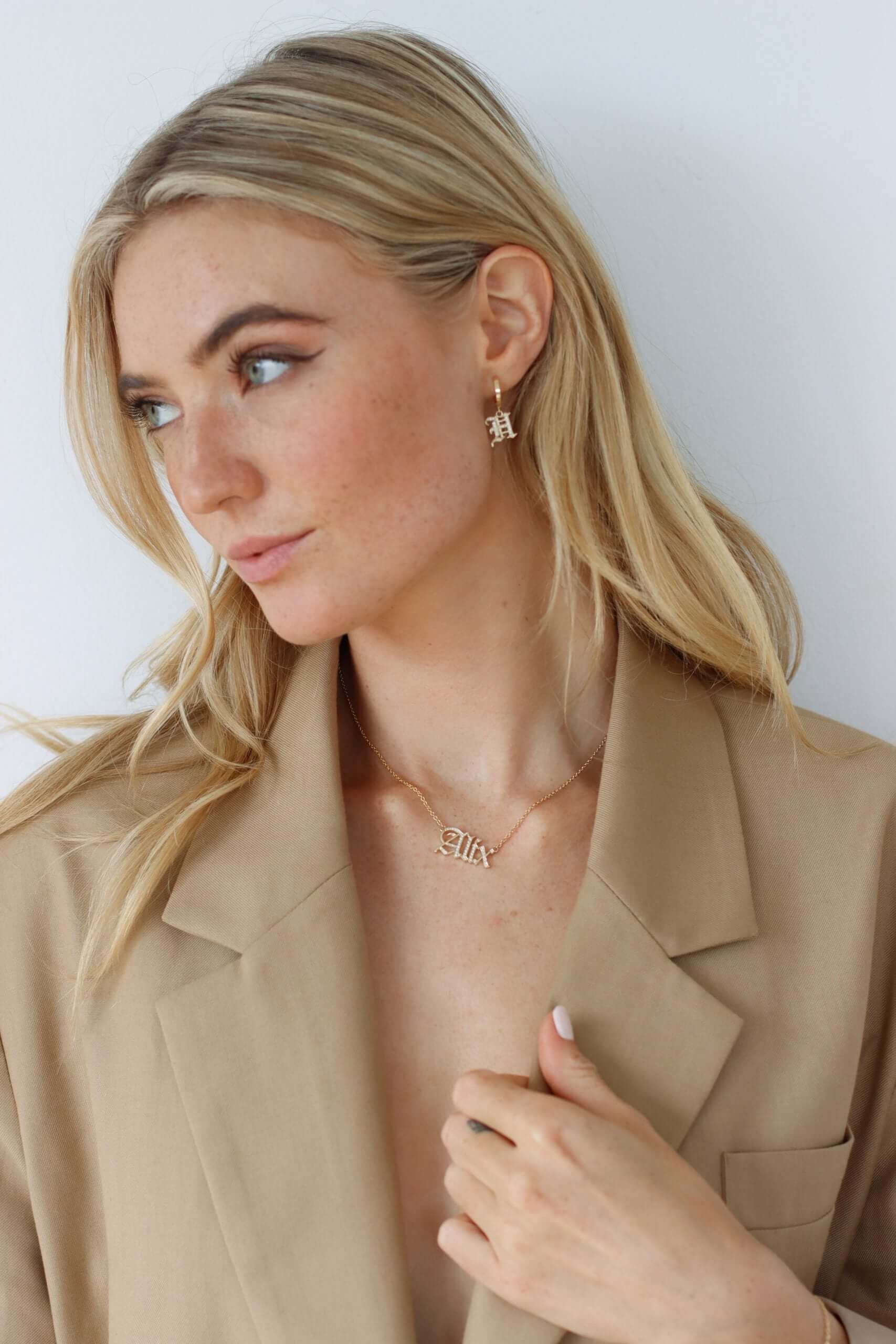 A woman with blonde hair wearing a tan blazer looks to the side. She wears the Test - Small Old English Initial Huggies, delicate earrings that add a subtle touch to her look. A gold necklace with a pendant rests against her neck as she softly holds it, posing elegantly against a light background.