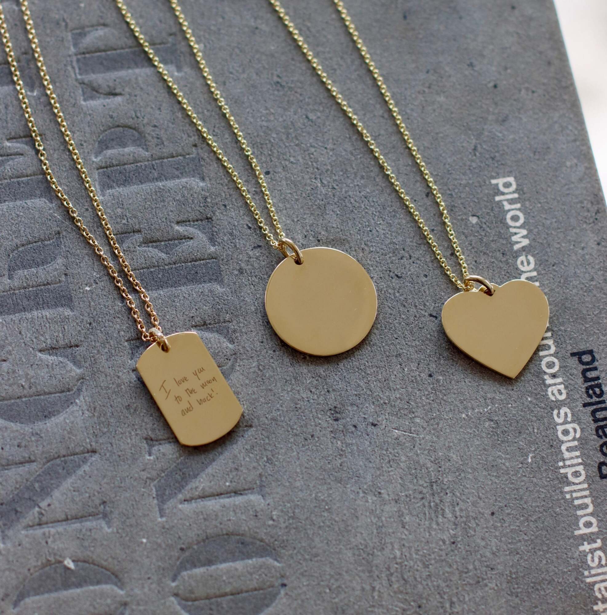 Three gold necklaces on a gray textured surface: one personalized keepsake with a rectangular pendant featuring engraved text, one Large Round Engravable Pendant Necklace, and one with a heart-shaped pendant—each piece a timeless example of engraved jewelry.