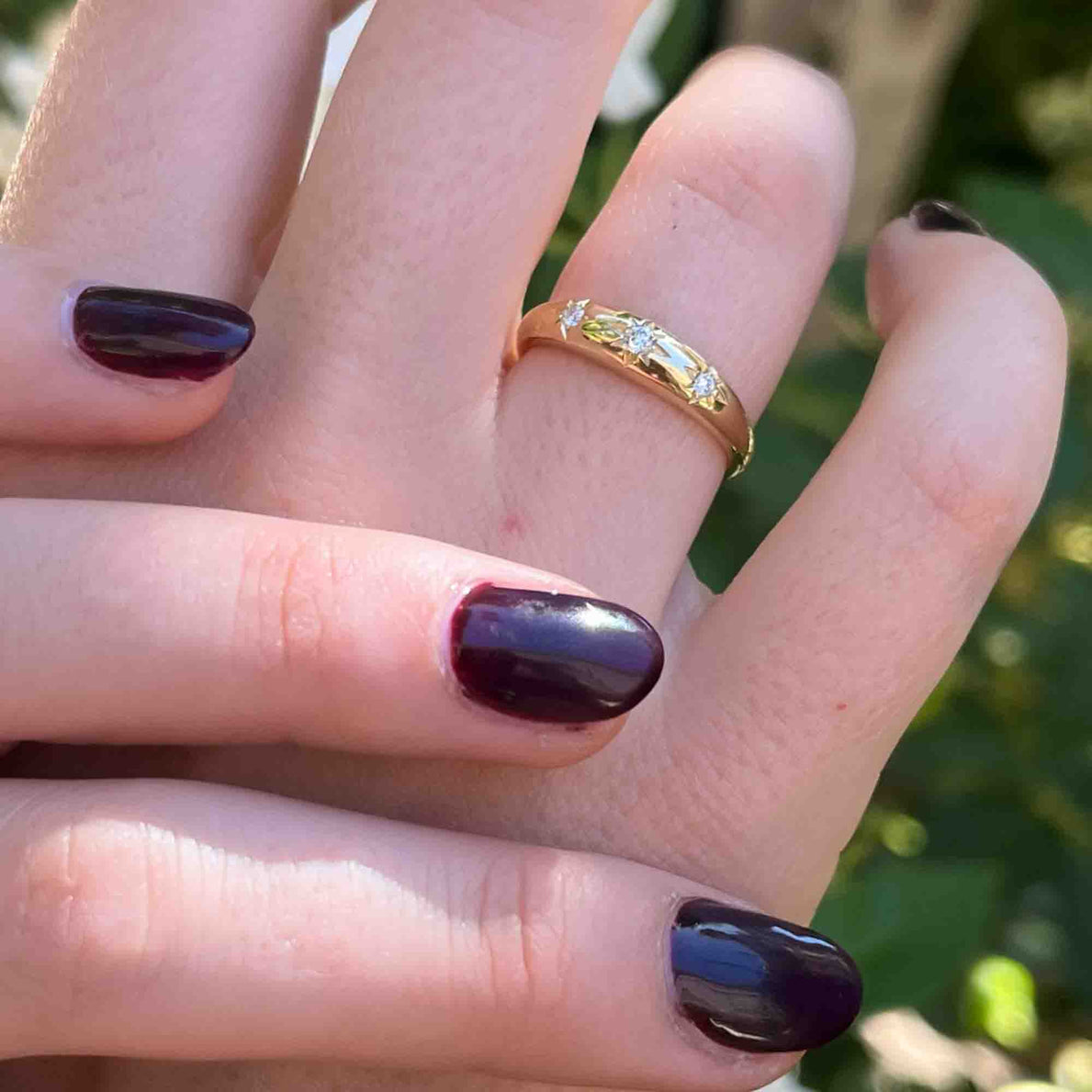 Two hands with dark nails, one above the other, display elegant jewelry. The top hand features a Diamond Star Dome Ring that sparkles amid the blurred greenery background. Preorder now to add this stunning accessory to your collection.