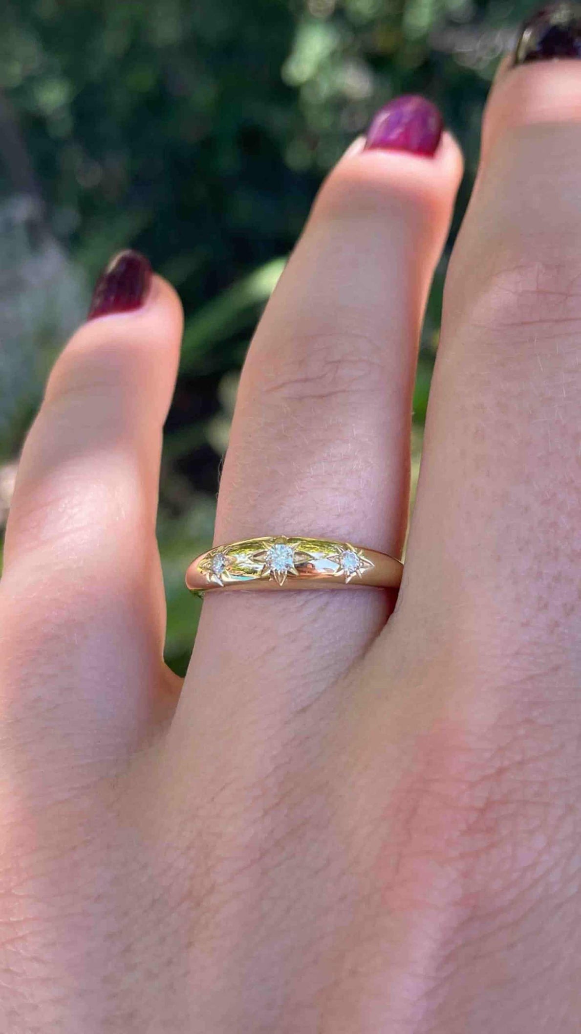 A hand with dark red nail polish elegantly displays the Diamond Star Dome Ring, featuring three gemstones in star-shaped settings. A blurred green foliage background highlights this exquisite jewelry. Available for preorder now.