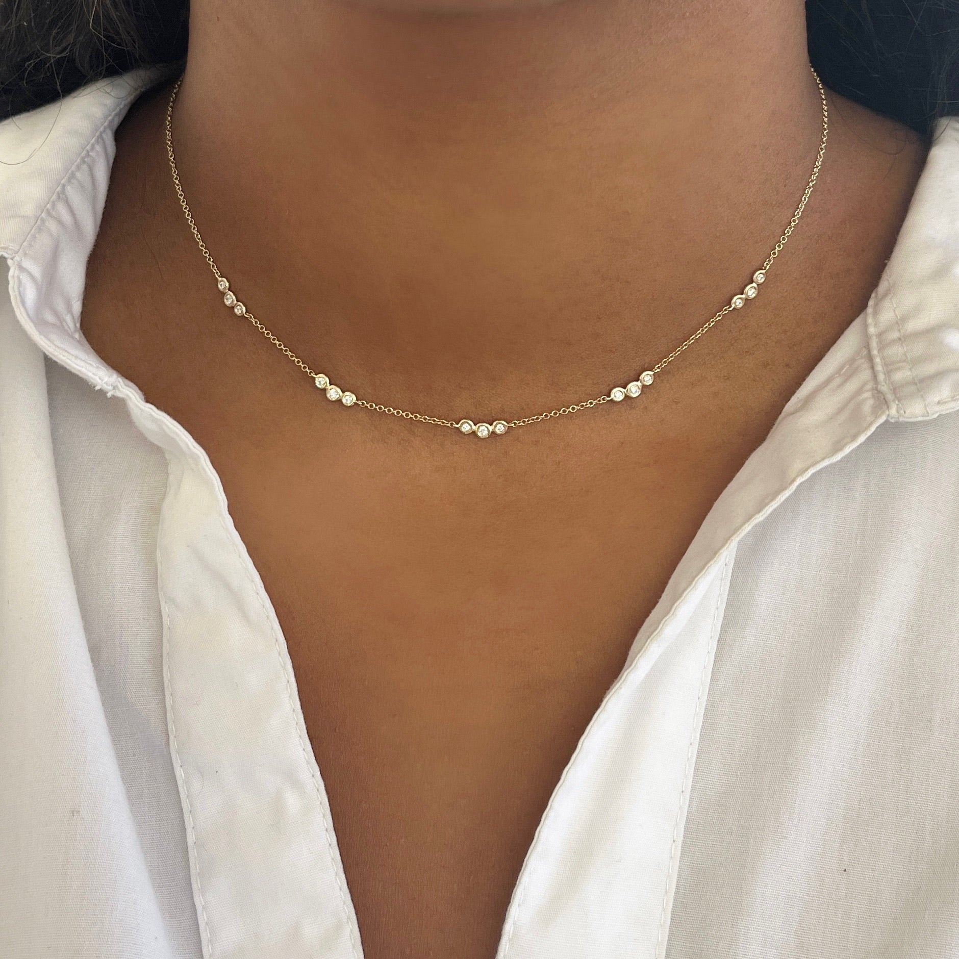 A person wearing the Bezel Diamond Trio Station Necklace from Janet Heller Fine Jewelry—a delicate gold chain adorned with trio diamond stations—paired with a white collared shirt.