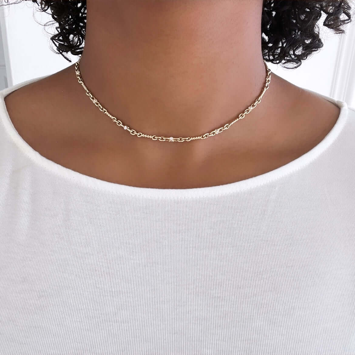 A close-up shows a person wearing the Diamond Belle Necklace by JanetHellerFineJewelry against a plain white shirt, with a softly blurred background. The chain of small linked loops rests lightly on their warm-toned skin, complementing their curly hair.