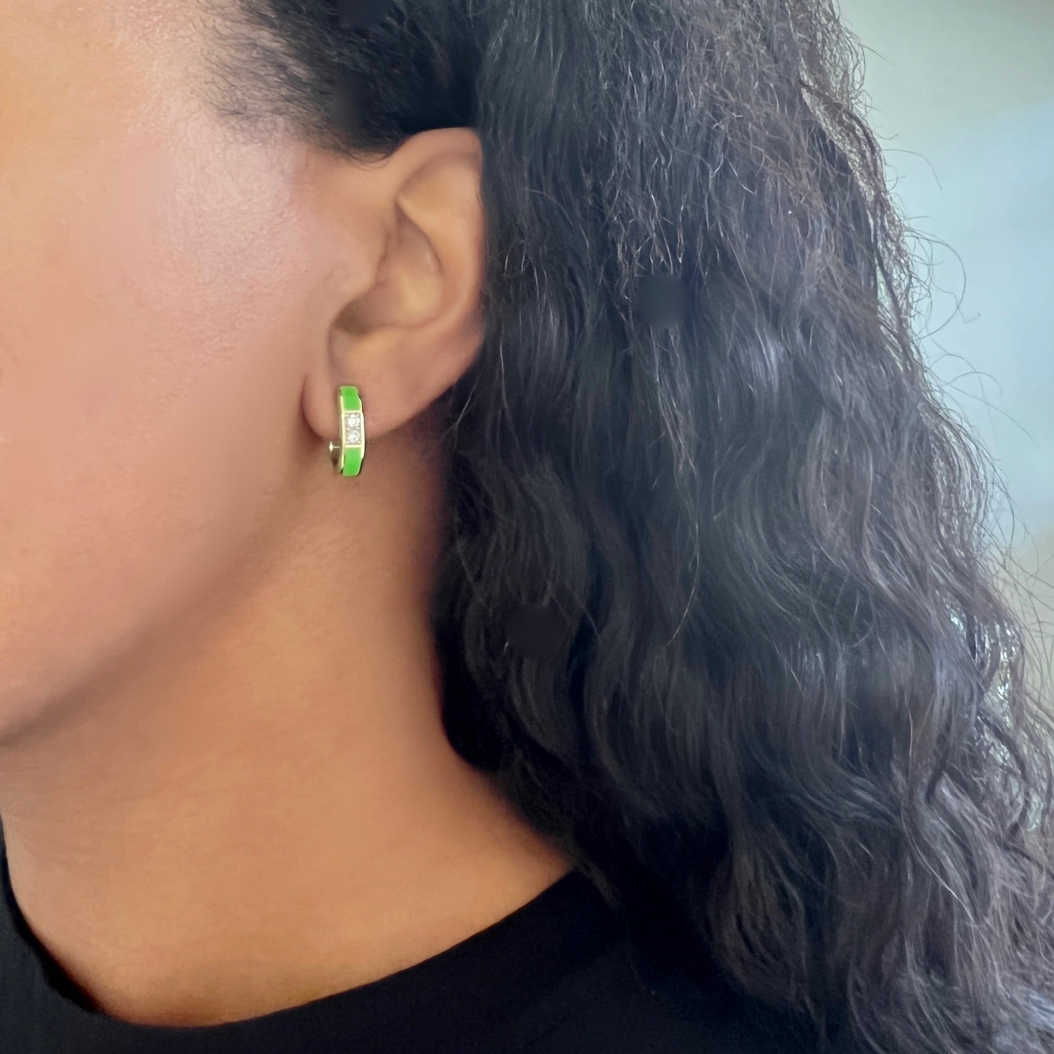 A person with long, wavy dark hair wears the U Shaped Green Enamel Hoops—small green earrings accented with silver, designed in the elegant style of Janet Heller. The photo captures a close-up side view of their ear and jawline.