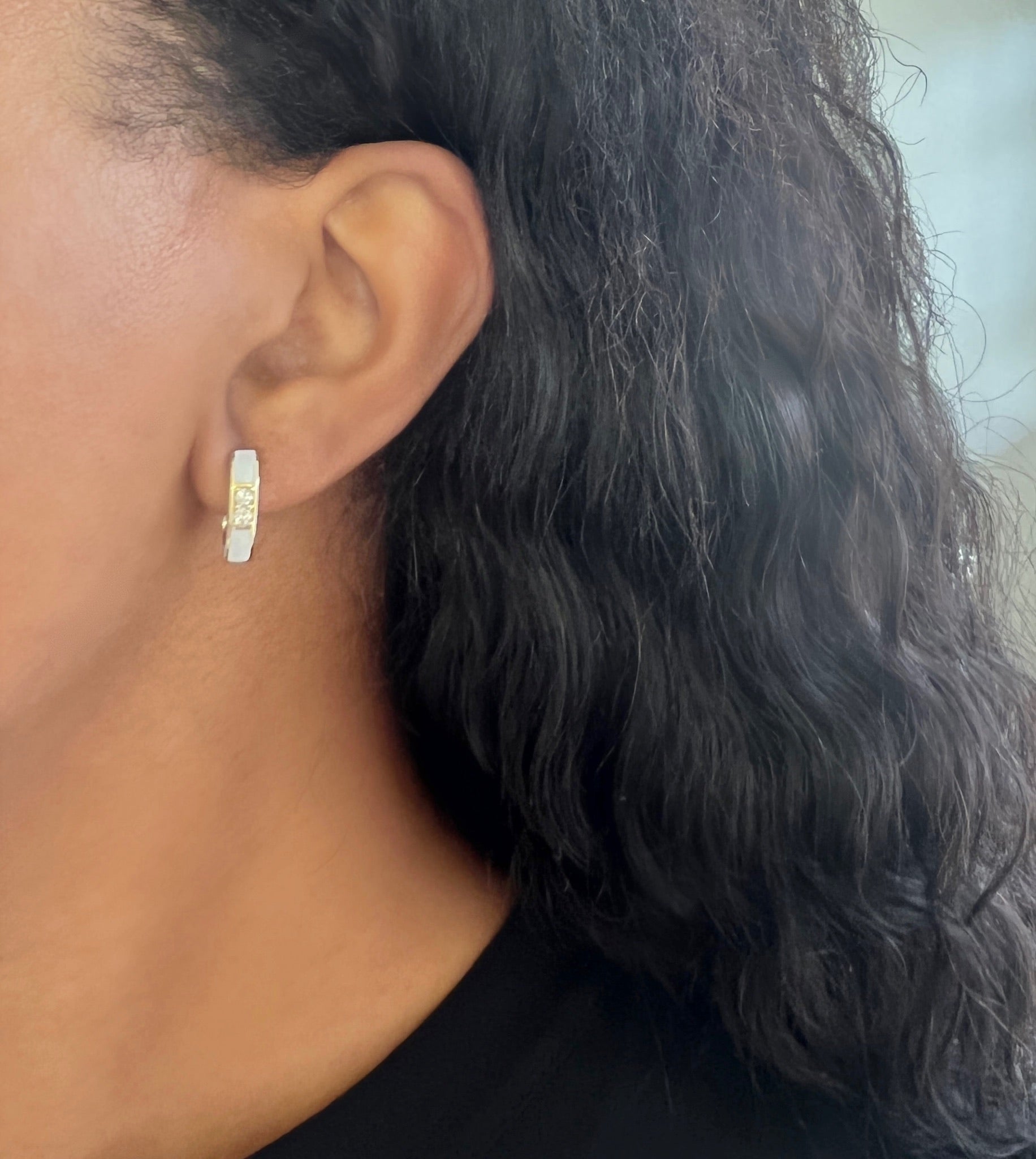 Close-up of a person's ear wearing the U Shaped White Enamel Hoops from Janet Heller fine jewelry—a small, rectangular hoop earring with a textured design in silver or white gold. The person has curly, dark hair and is dressed in a black top. For jewelry inquiries or custom options, please reach out for more information.