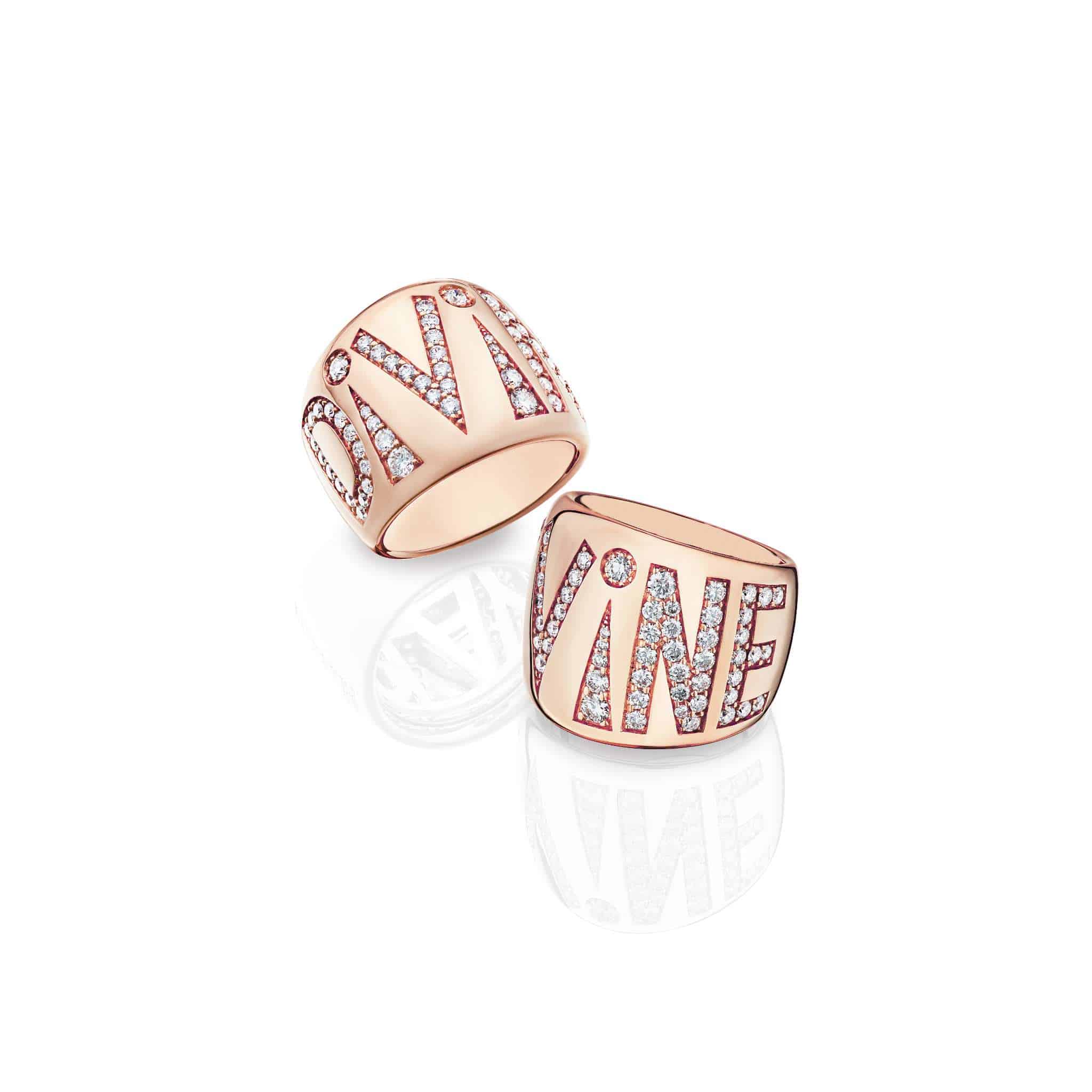 Two Divine Play Rings in rose gold, each with the word DIVINE spelled out in capital letters and encrusted with small diamonds, rest on a white surface with their reflections visible.