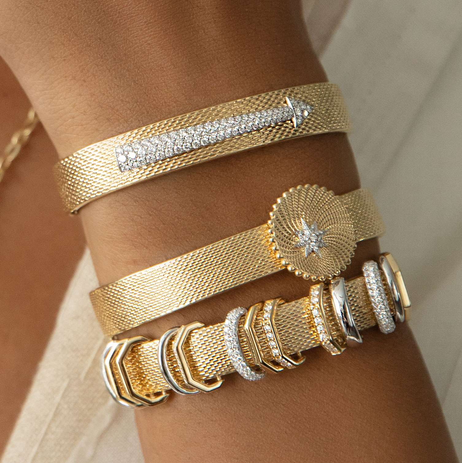A close-up of a wrist wearing three gold bracelets, including the Diamond Arrow Bangle featuring a row of diamonds and arrow detail, alongside a bracelet with a round sunburst design and diamond, and another with stacked textured and diamond-accented rings.