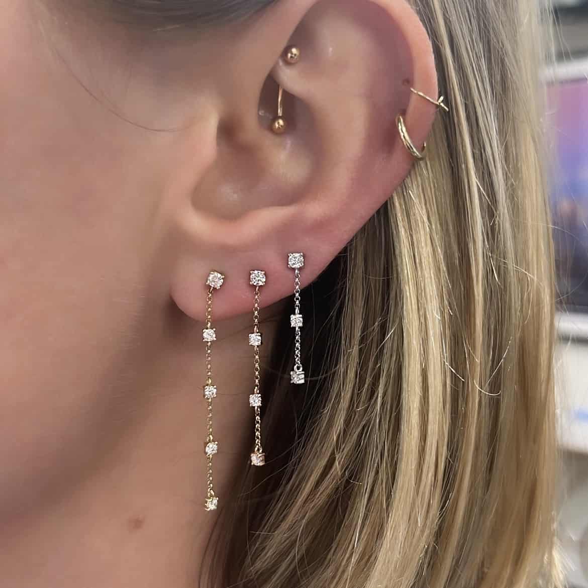 A close-up of an ear with multiple piercings, showcasing the elegant 3 Drop Diamond Finley Earrings and two gold hoops in the upper ear. The person has blonde hair and is displaying fine jewelry adorned with sparkling gems.