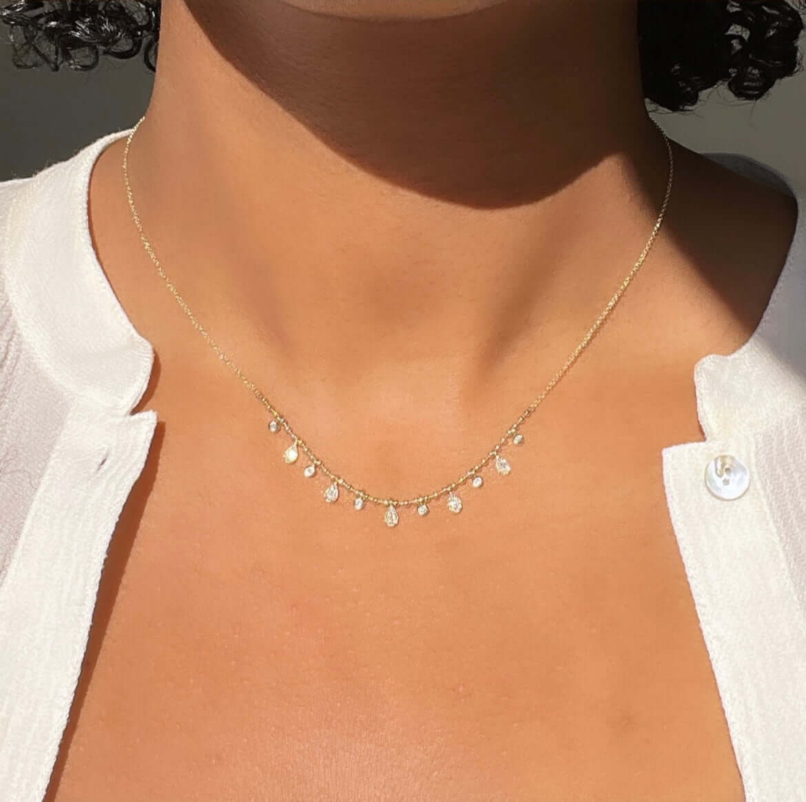 A person elegantly wears the Beaded Bezel & Pear Diamond Necklace with customizable charms. Against a light-skinned neck, it beautifully complements a white button-down shirt. The softly lit background enhances the scenes serene sophistication.