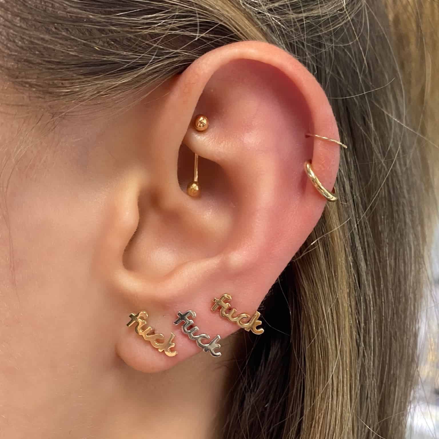 A close-up of an ear with multiple piercings displays three gold Mini Plain F**K Stud earrings—a striking custom design—paired with a gold hoop in the upper ear and a barbell in the inner ear, all embodying the edgy elegance of Janet Heller fine jewelry.