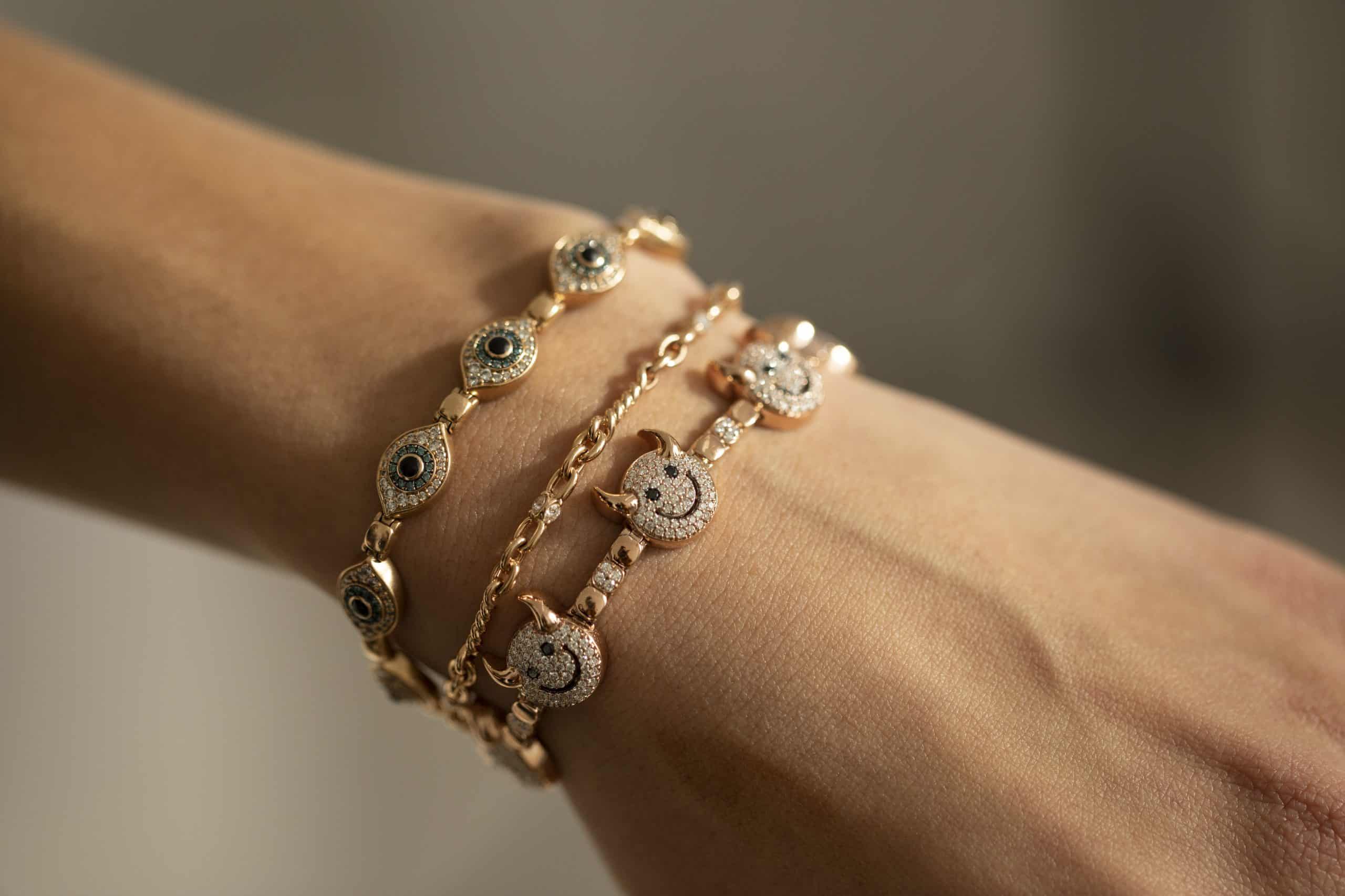 A close-up of a wrist adorned with two gold bracelets, including the Lucky Devil Bracelet (SKU: BRA-1213-Y), which is embellished with circular blue and white stones, paired with another bracelet featuring delicate round diamond-encrusted charms. This exquisite jewelry piece is elegantly displayed against a softly blurred background, capturing an aura of sophistication and good fortune.