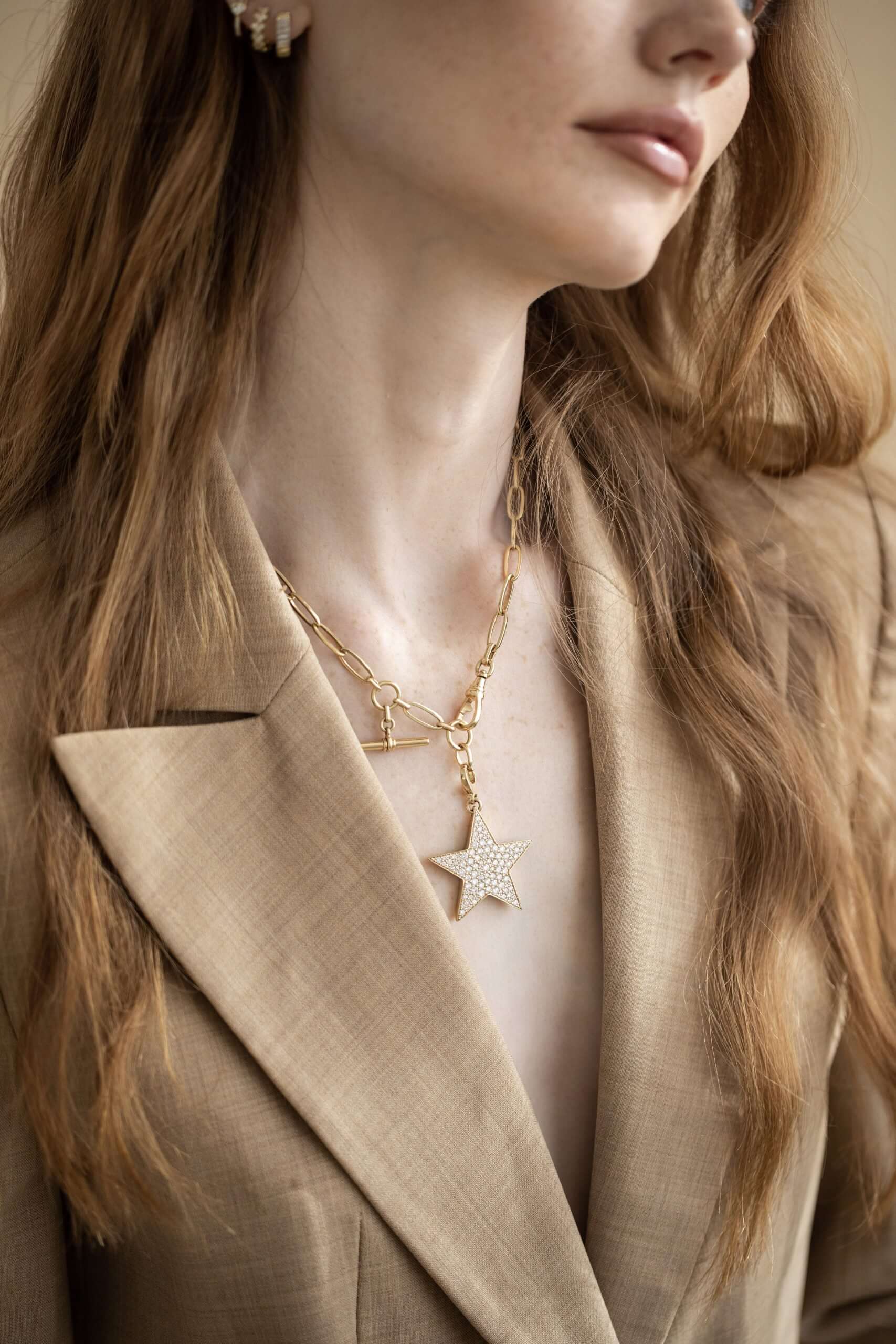 A woman with long, wavy, light brown hair wears a tan blazer and layered gold necklaces from Janet Heller Fine Jewelry, including the Florence Chain, one of which features a delicate star pendant among other chic charms. Only her lower face and torso are visible.