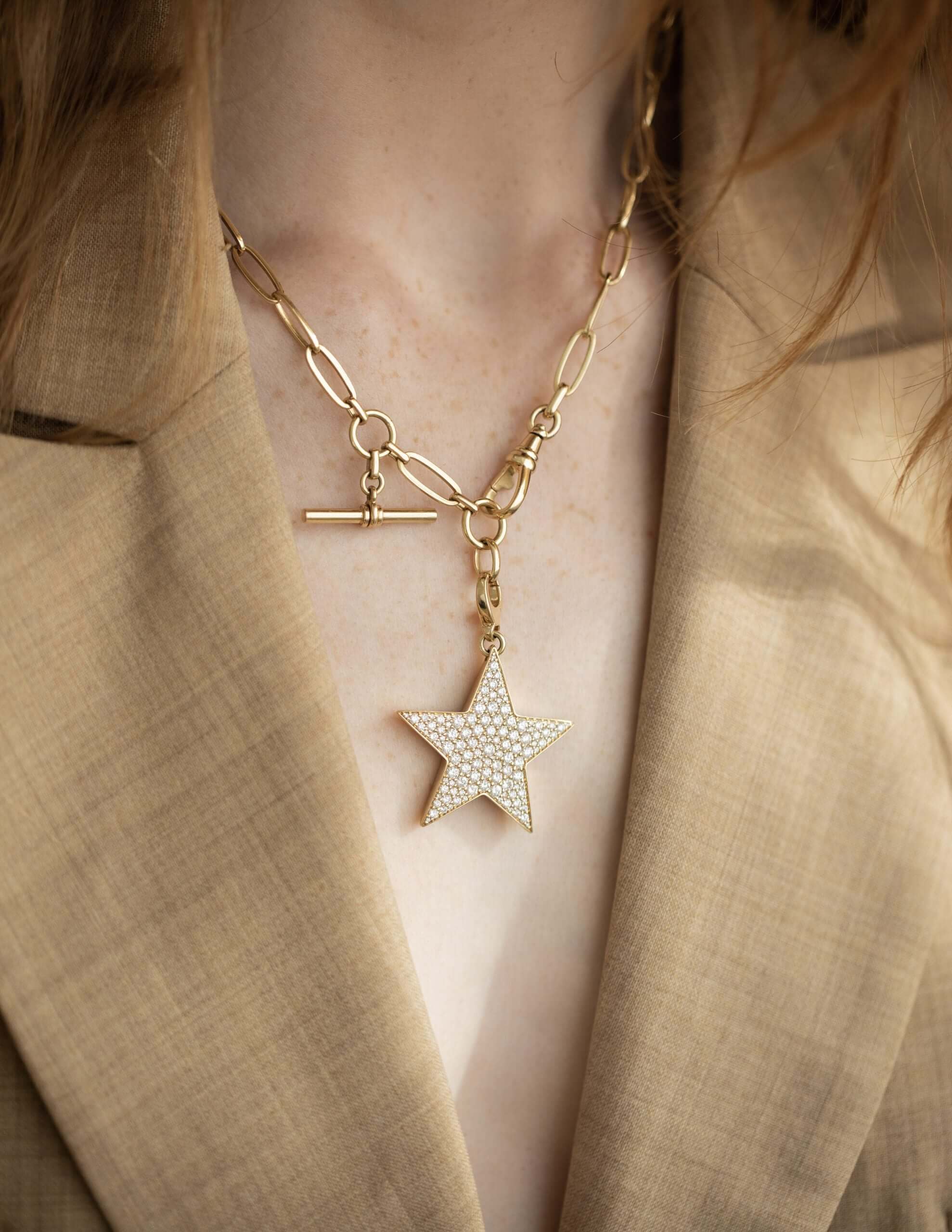 A person wearing a tan blazer and the Florence Chain from Janet Heller Fine Jewelry, featuring a chunky gold design with a large, rhinestone-studded star pendant adorned with elegant charms.