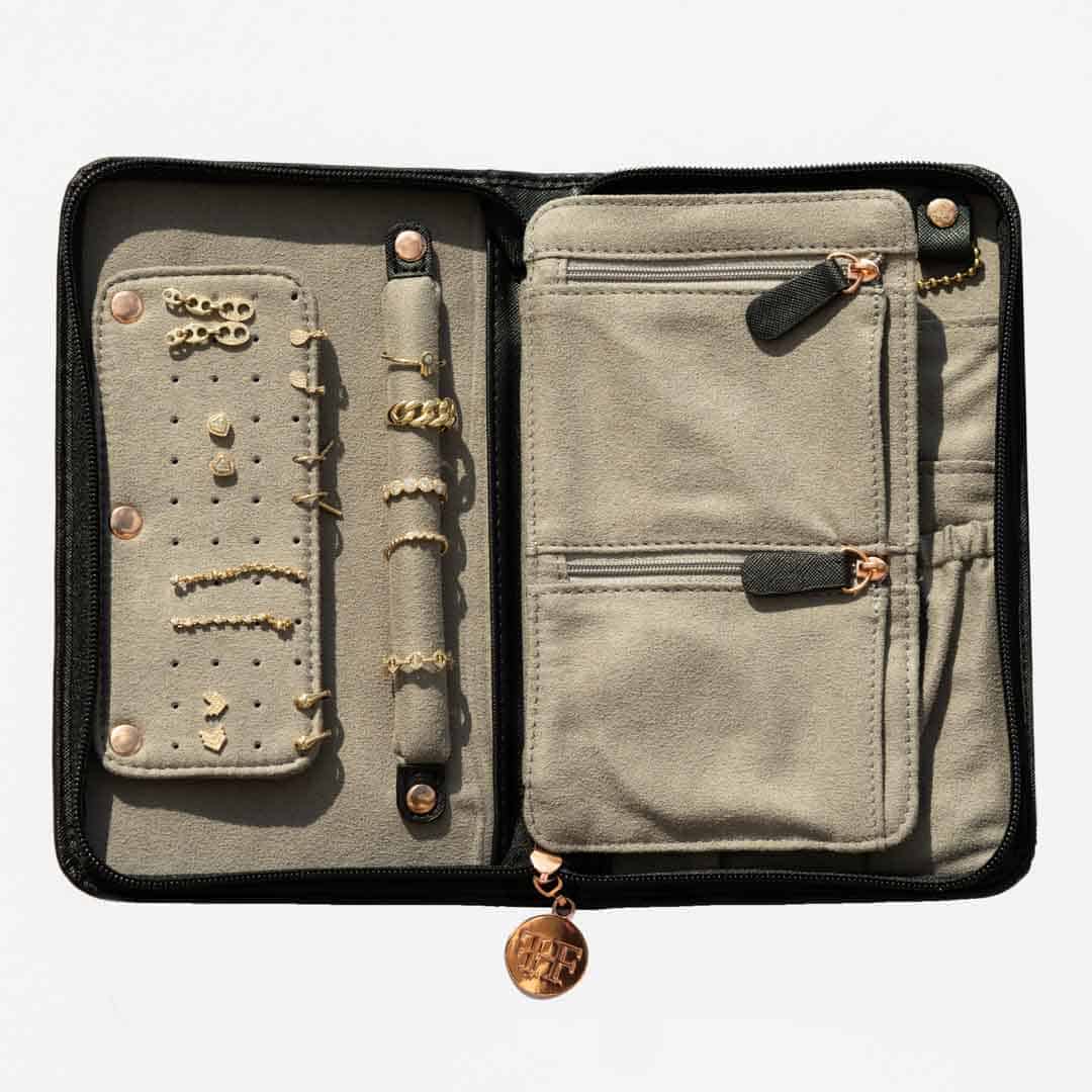 A beige vegan leather Travel Case with multiple compartments, zippers, and snap closures stores gold rings, necklaces, earrings, and a bracelet. This elegant organizer for jewelry features a round zipper pull with initials displayed at the bottom.