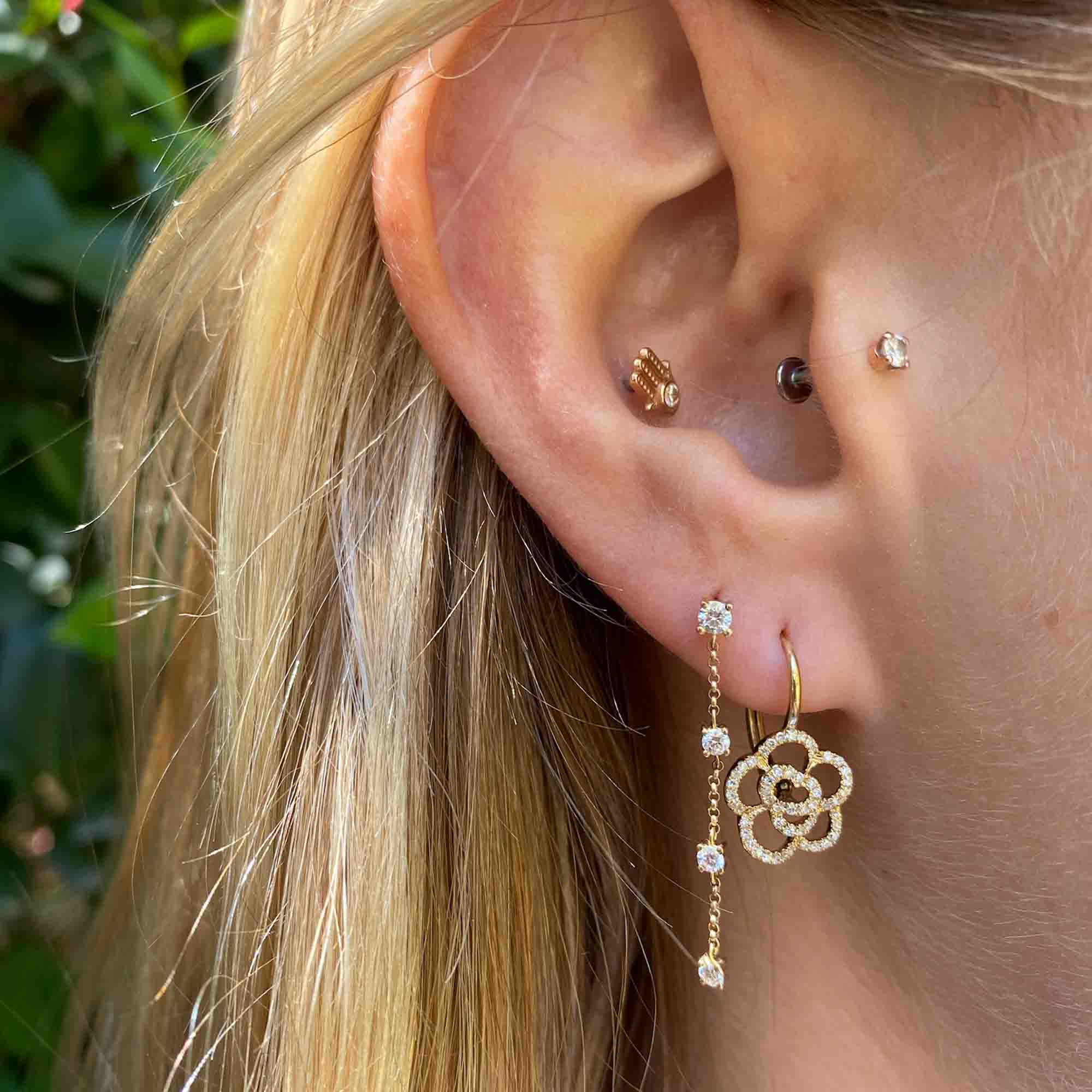 Here is your rewritten sentence using the provided product data: Close-up of a woman's ear with five piercings, showcasing fine jewelry including gold and 4 Drop Diamond Finley Earrings, as well as a dangling floral drop earring. Blonde hair is visible, set against a backdrop of softly blurred greenery.