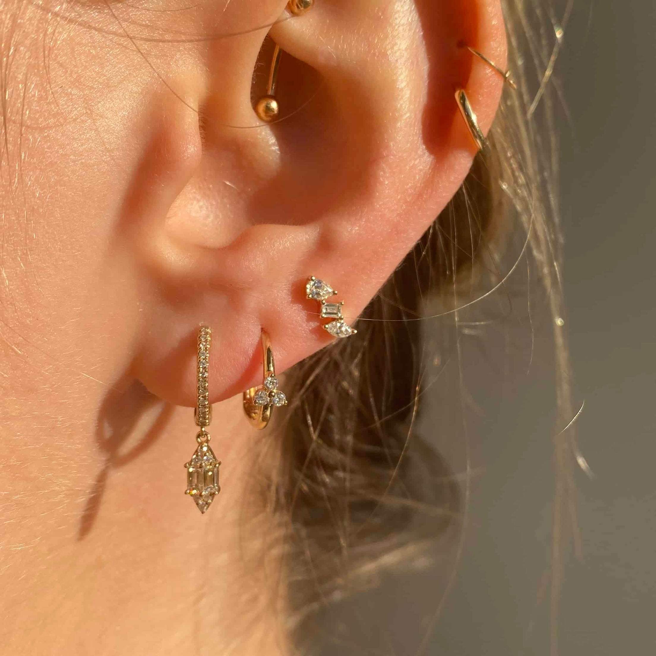 A person wears elegant jewelry: Diamond Jolie Huggies with a geometric charm, a stone cluster earring, and a simple cartilage hoop. The blurred background enhances elegance. Contact us for inquiries or customizations.