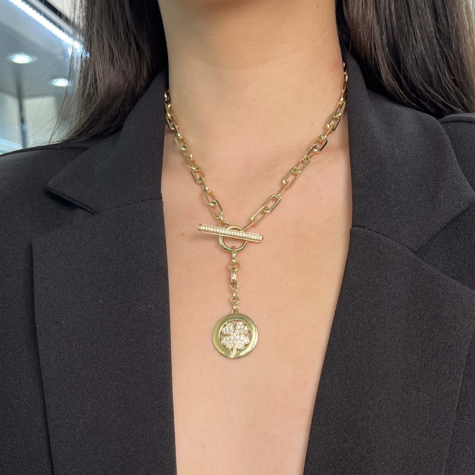 A person in a black blazer is featured wearing Janet Heller Fine Jewelrys Diamond Clover Disc Charm necklace, which has an intricate medallion, toggle clasp, and gold chain. The softly blurred background highlights the bespoke charm and elegant attire.
