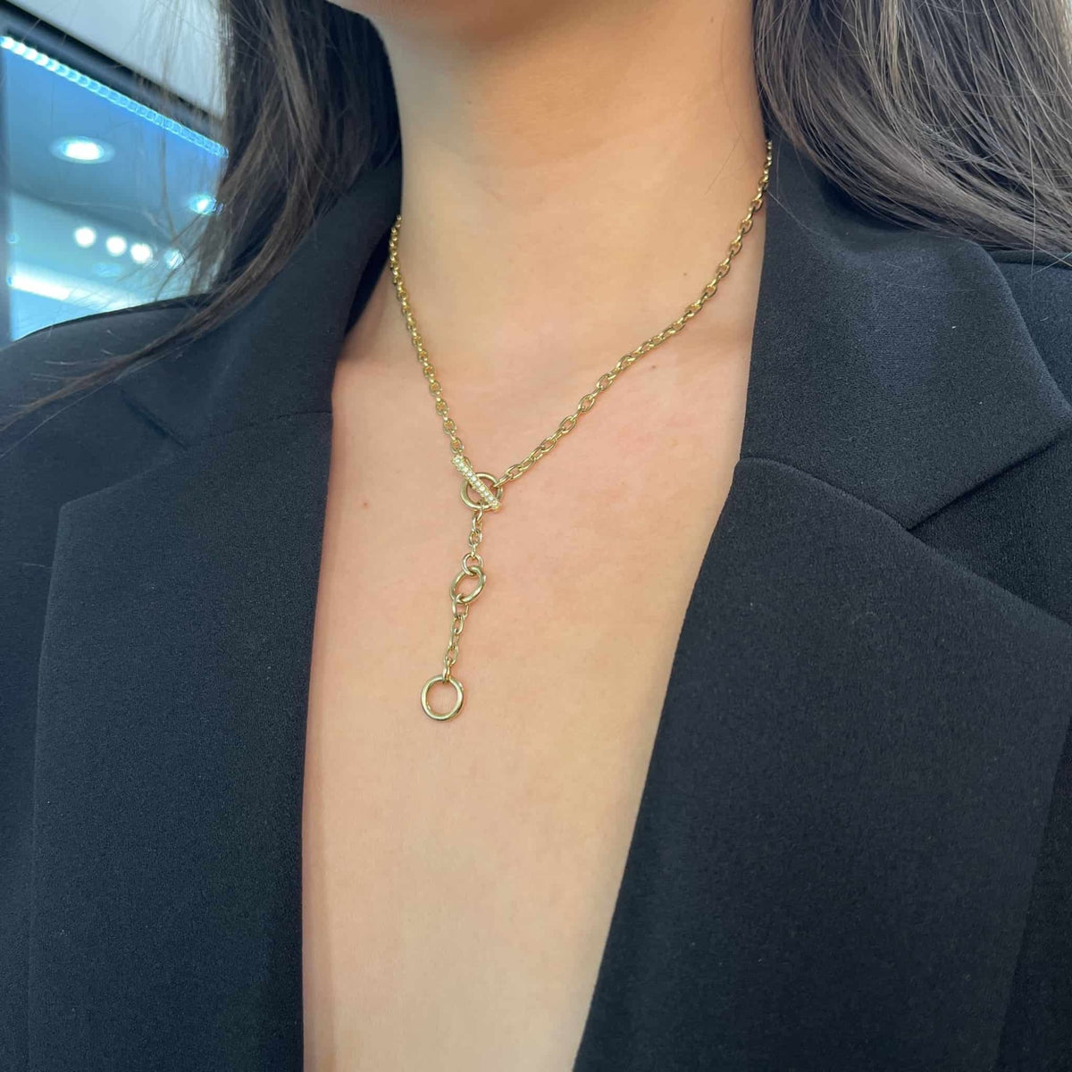 A person in a black blazer with an open neckline wears the Janet Heller Fine Jewelry London Chain gold necklace, adorned with two circular charms. The background is softly blurred with blue and white lights.