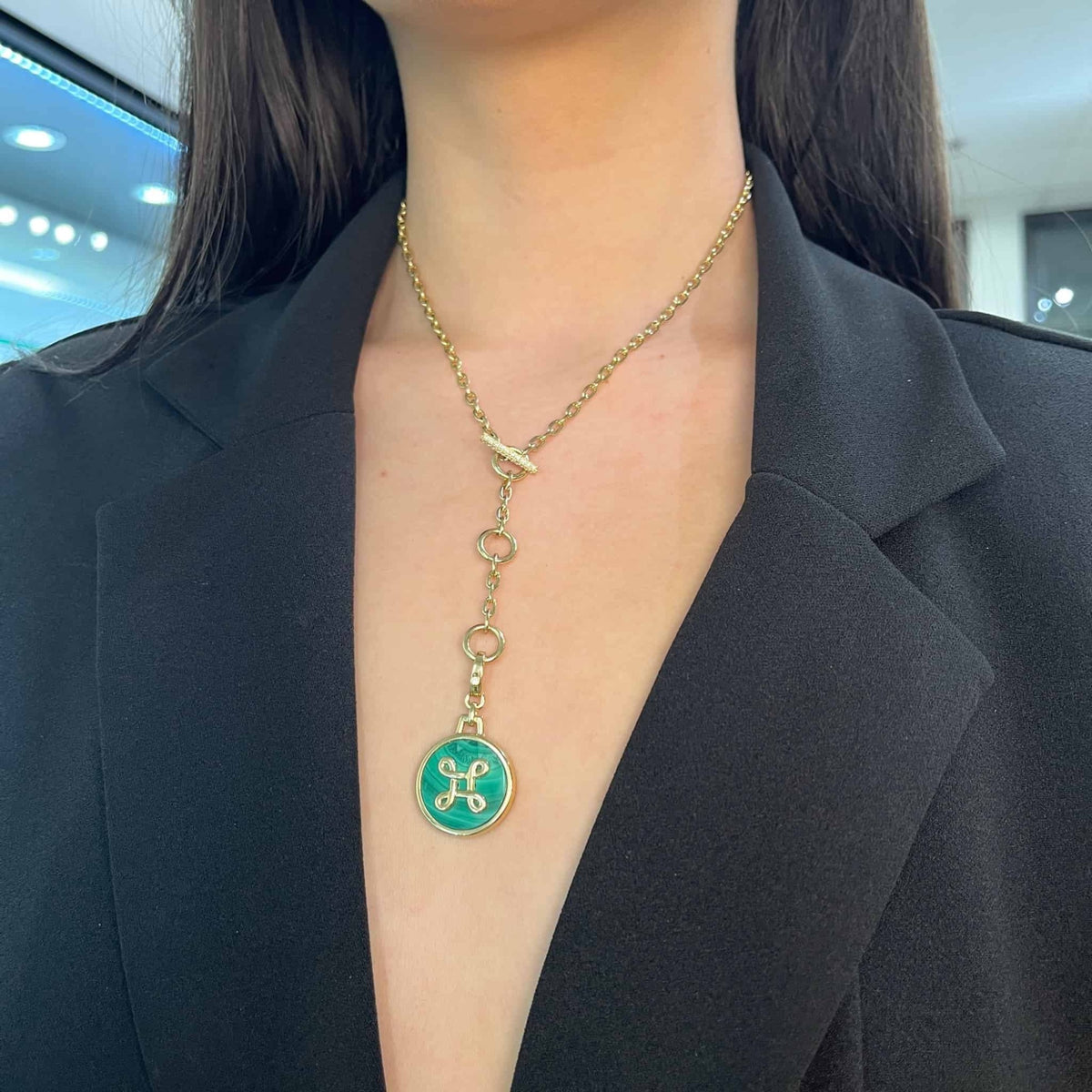 Close-up of a person wearing a black blazer and a gold necklace, the London Chain from Janet Heller Fine Jewelry, showcasing a unique green pendant with decorative golden design. Its elegant charm is enhanced by softly lit shelves and lights reflecting in the background.