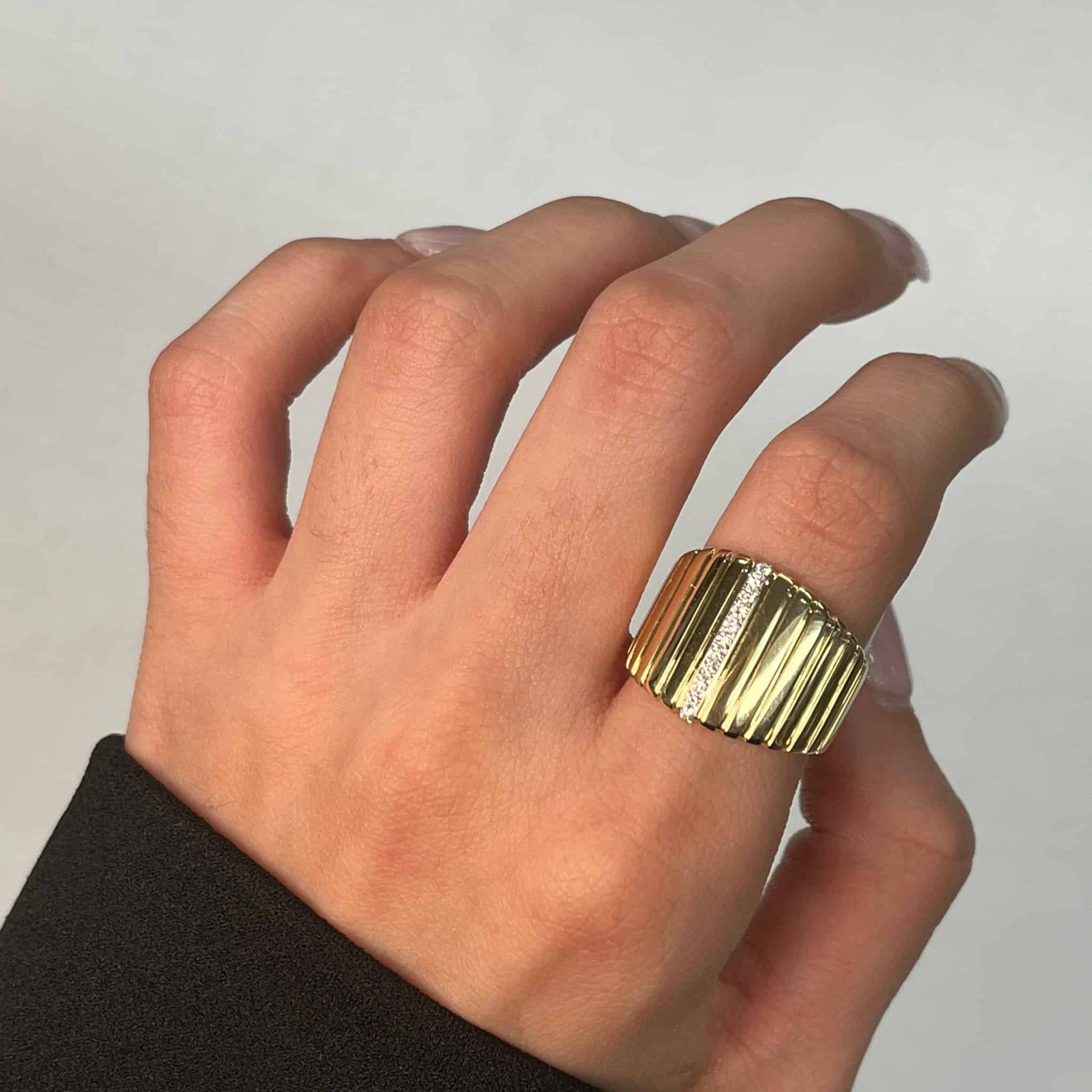 A hand adorned with the Large Diamond Ridge Ring—a gold ribbed ring—is showcased against a neutral backdrop. The wearer, in a black long-sleeve, highlights the rings textured design and lustrous finish, making it an exquisite item available for preorder.