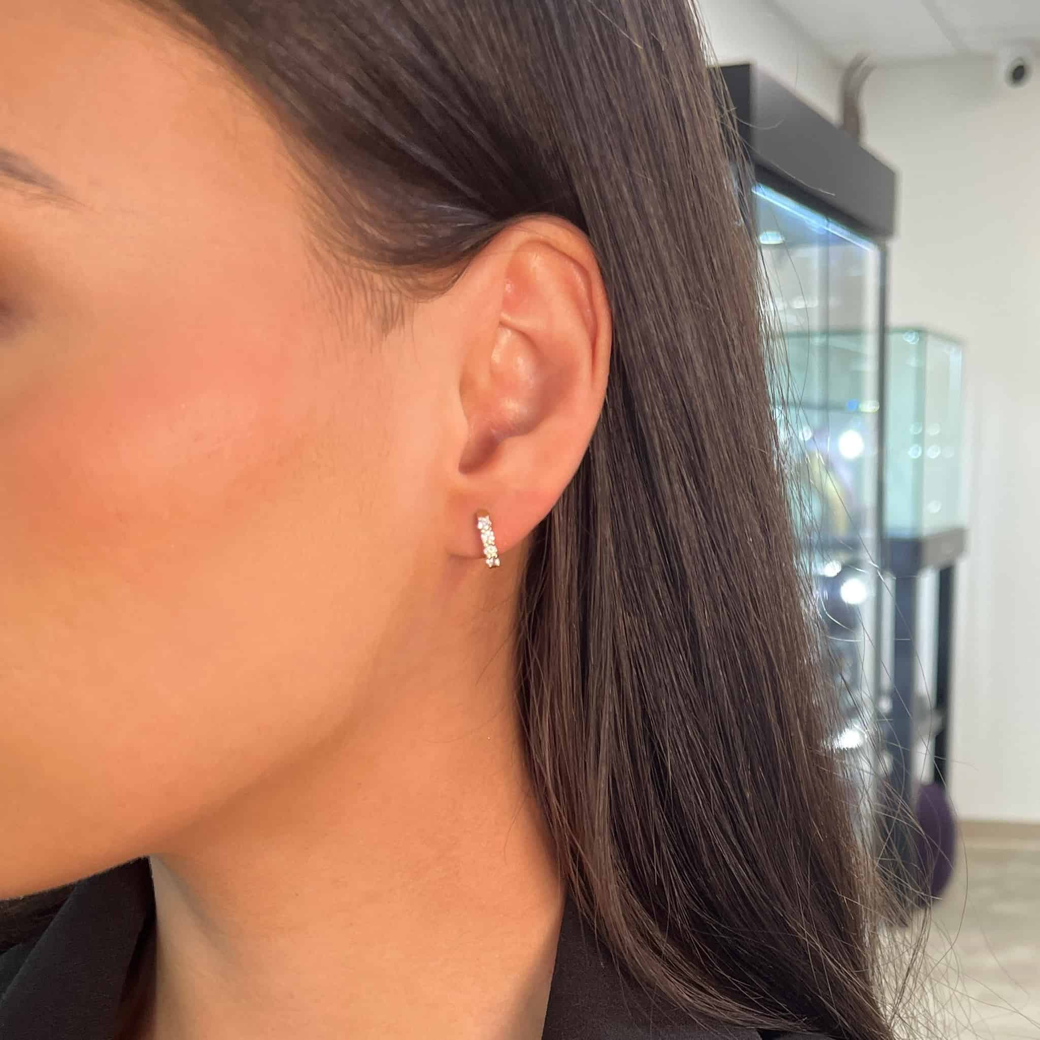 A close-up view of a woman’s ear adorned with the Diamond Dixie Huggies ($1,125.00, SKU: EAR-540-W) from Janet Heller Fine Jewelry. She has straight, dark hair and appears to be indoors, possibly at a boutique specializing in fine jewelry. The small gold huggie earring features embedded diamonds for an elegant touch.