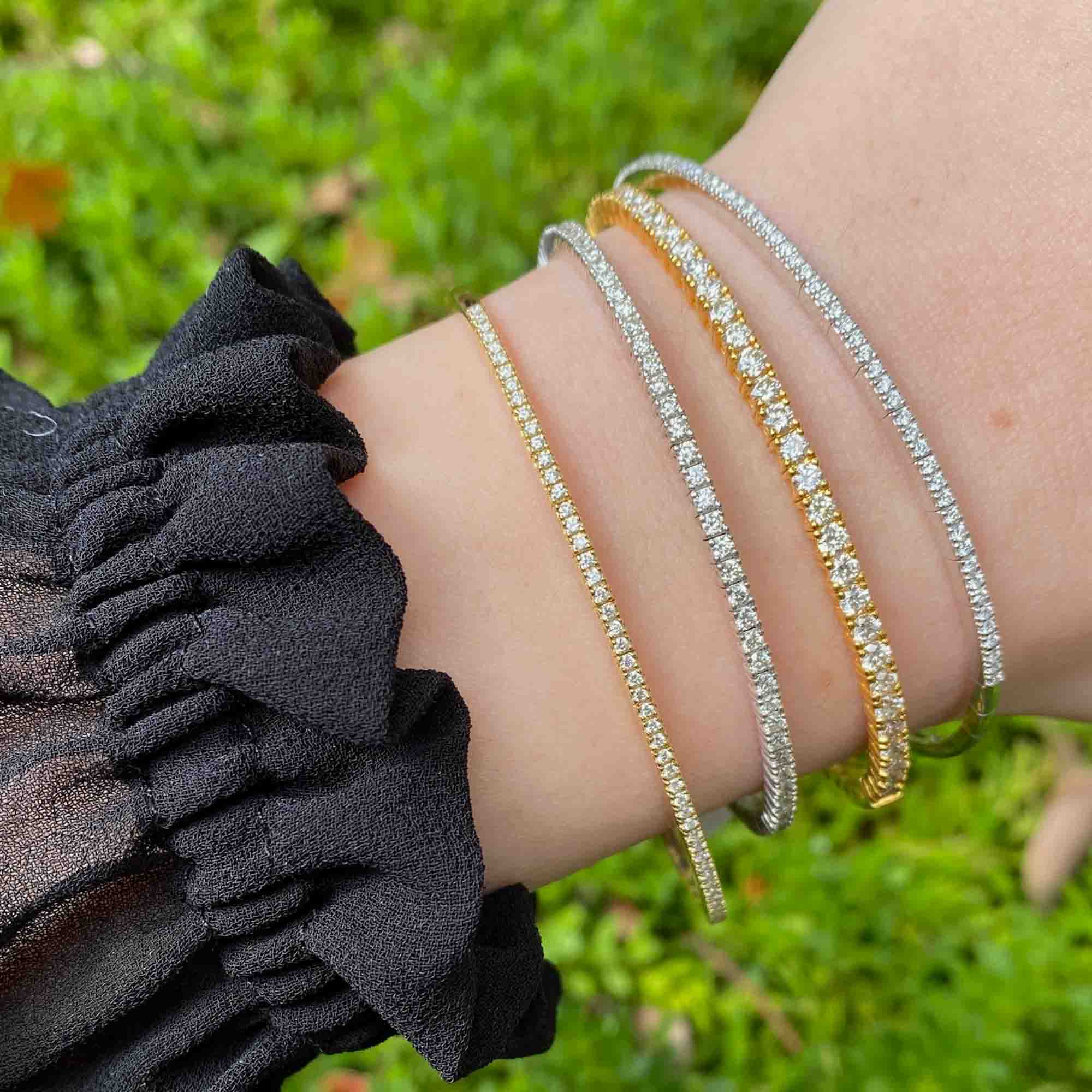 A close-up of a wrist adorned with four sparkling Diamond London Bangles from Janet Heller Fine Jewelry—two in gold and two in silver—shining beautifully above a ruffled black sleeve. The fine jewelry catches the light against the lush green grass backdrop.