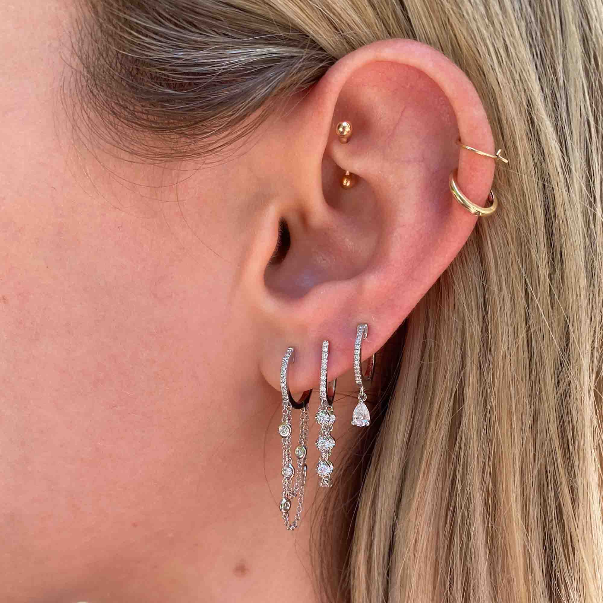 A close-up of a woman's ear showcases exquisite fine jewelry: three dangling diamond earrings, a small stud, and two gold hoop piercings on the upper ear. She has blonde hair. The elegant arrangement highlights the artistry of custom jewelry often seen in Janet Heller Fine Jewelry collections, featuring the Diamond Loop Huggies.