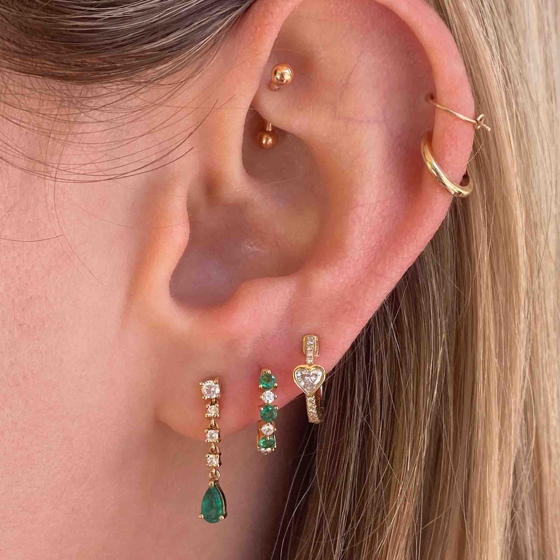 A close-up showcases an ear adorned with fine jewelry: a gold hoop in the upper lobe, a small stud in the cartilage, another hoop around the earlobe, and two Pear Emerald Drop Earrings. Perfect for customization or product inquiries.