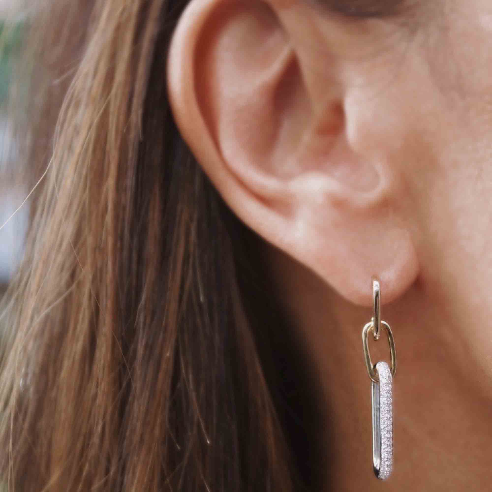 Close-up of a person’s ear with straight brown hair, wearing the Two Tone Diamond Paperclip Earrings—gold earrings featuring two linked oval shapes, one of which is encrusted with small sparkling stones.