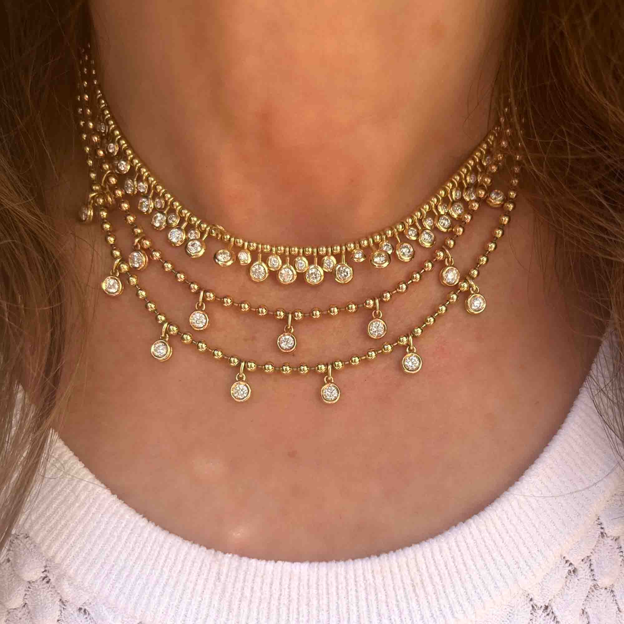 Certainly! Here is the revised sentence using your product data: A close-up of a person wearing the Diamond Kinsley Necklace from Janet Heller Fine Jewelry, a layered gold necklace adorned with small round pendants, each set with a clear gemstone. Paired with a white textured top, this piece is perfect for those seeking custom jewelry or who have jewelry inquiries.